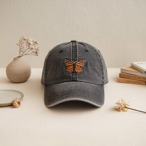 Puede incluir: Gorra de béisbol gris carbón con acabado desgastado. Cuenta con un parche bordado de mariposa en naranja y negro. El gorro es de diseño clásico de seis paneles con visera curva.