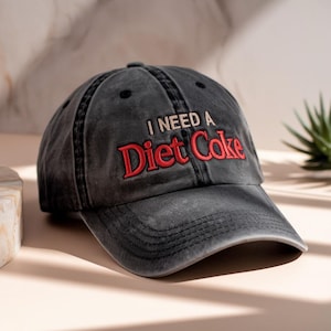 May include: A faded black baseball cap featuring the embroidered phrase "I NEED A Diet Coke" in red. The cap has a curved peak and a traditional six-panel construction, ideal for everyday use.