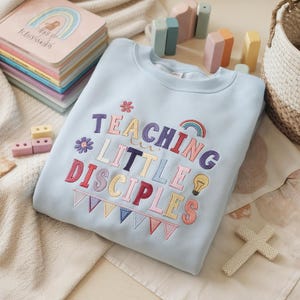 Könnte beinhalten: Hellblaues Sweatshirt mit dem gestickten Text "TEACHING LITTLE DISCIPLES" in bunten Buchstaben. Das Design umfasst einen Regenbogen, eine Blume, eine Glühbirne und Wimpel. Kinderspielzeug ist um das Sweatshirt herum angeordnet.