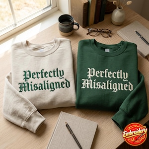 May include: Two crewneck sweatshirts, one cream with dark green embroidery and one dark green with cream embroidery. Both feature the text "Perfectly Misaligned" in a gothic-style font across the chest, with long sleeves and ribbed cuffs.