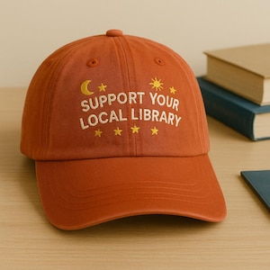 Peut inclure: Casquette de baseball orange brûlé avec le texte "SUPPORT YOUR LOCAL LIBRARY" brodé en blanc. Le design comprend un croissant de lune, un soleil et des étoiles. La casquette est en tissu doux et possède une visière incurvée.