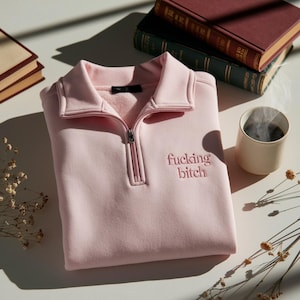 Pode incluir: Um moletom rosa claro com gola com zíper e o texto bordado "fucking bitch". O moletom está dobrado sobre uma superfície branca, com livros e uma xícara de café ao fundo.