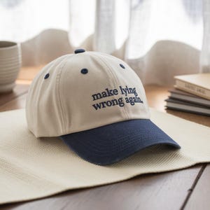 May include: A beige and navy blue baseball cap with the phrase "make lying wrong again" embroidered in navy blue. The cap has a curved brim and a rounded crown with navy blue eyelets.