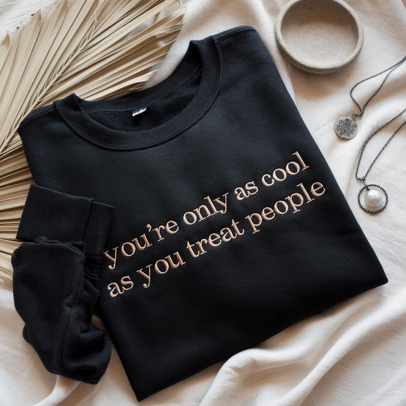 May include: Black sweatshirt with the embroidered text "You're only as cool as you treat people." The sweatshirt is displayed with a folded sleeve, a small bowl, and two necklaces.
