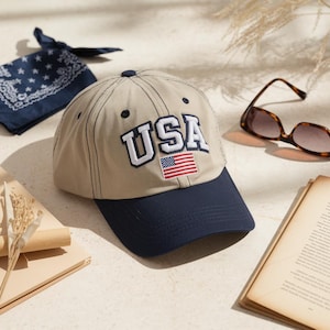 Könnte beinhalten: Beige Baseball Cap mit marineblauem Schirm, mit dem Wort "USA" in weißen Buchstaben und einer kleinen amerikanischen Flagge. Ein marineblaues Bandana und eine Schildpatt-Sonnenbrille sind ebenfalls zu sehen.