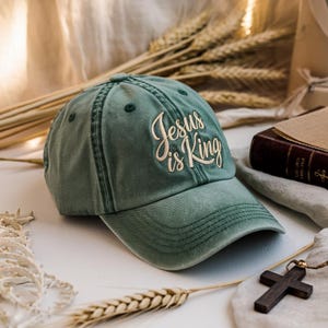 Könnte beinhalten: Eine salbeigrüne Baseballkappe mit dem gestickten Text "Jesus is King" in kursiver Schrift. Die Kappe besteht aus einem weichen, abgenutzten Material und hat einen gebogenen Schirm. Die Kappe ist ein religiöser Artikel.