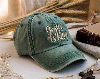 May include: A sage green baseball cap with the embroidered text "Jesus is King" in a cursive font. The cap is made of a soft, worn material and has a curved brim. The cap is a religious item.