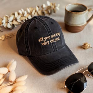 Könnte beinhalten: Eine schwarze, ausgewaschene Baseballkappe mit dem gestickten Text "eff you see kay why oh you" in Hellbraun. Die Kappe befindet sich neben einer Kaffeetasse und einer Sonnenbrille.