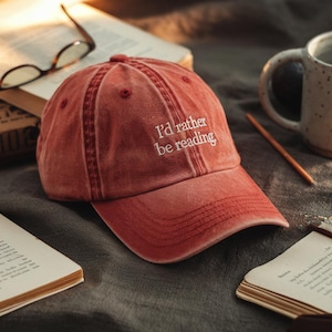 May include: A faded red baseball cap with the embroidered text "I'd rather be reading" in white. The cap is sitting on a dark gray surface with open books, glasses, and a mug in the background.