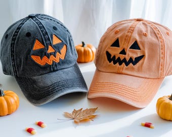 May include: Two baseball caps featuring jack-o'-lantern designs. One is faded black, the other is orange. Both have orange and black embroidered details. Small pumpkins and candy corn are scattered around the hats, suggesting a Halloween theme.