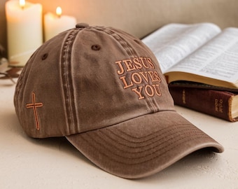 以下が含まれることがあります： オレンジ色の「JESUS LOVES YOU」の刺繍が入った茶色の野球帽。側面に十字架が刺繍されています。帽子は柔らかく、使い古された素材で作られています。