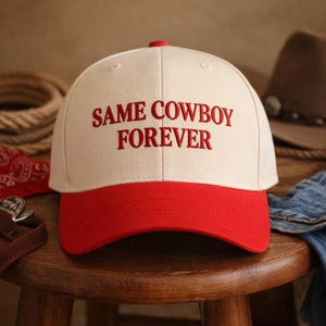 Può includere: Un cappellino da baseball con corona color crema e visiera rossa. La scritta "SAME COWBOY FOREVER" è ricamata in rosso sul davanti. Un bottone rosso è in cima. Il cappello è su uno sgabello di legno.