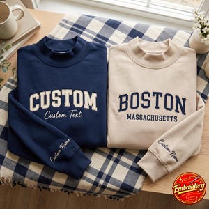 Puede incluir: Dos sudaderas de cuello alto con letras bordadas. La sudadera azul marino muestra la palabra CUSTOM con Custom Text debajo y Custom Name en la manga. La sudadera beige muestra BOSTON MASSACHUSETTS y Custom Name en la manga.