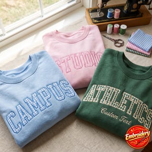 May include: Three crewneck sweatshirts in light blue, light pink, and dark green. The blue sweatshirt features the word CAMPUS, the pink one says STUDIO, and the green one says ATHLETICS and Custom Text, all in gingham-patterned applique lettering.