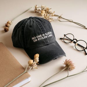 May include: A faded black baseball cap with the white text "NO ONE IS ILLEGAL ON STOLEN LAND". The cap is on a neutral surface with a notebook, round eyeglasses, and dried flowers.