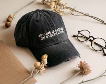 May include: A distressed black baseball cap with the text "NO ONE IS ILLEGAL ON STOLEN LAND" in white. The cap is placed on a neutral surface with a notebook, eyeglasses, and dried flowers.
