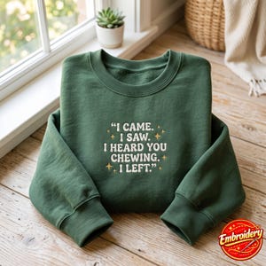 May include: A forest green crewneck sweatshirt featuring white embroidered text that reads, "I CAME. I SAW. I HEARD YOU CHEWING. I LEFT." The design is accented with small, gold-colored embroidered star shapes surrounding the central quote.