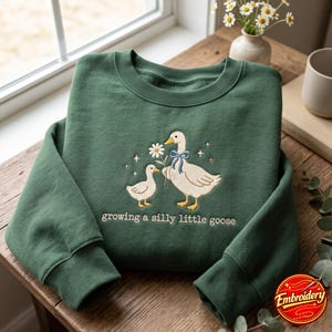 May include: A forest green crewneck sweatshirt featuring an embroidered design of a large goose with a blue ribbon and a small gosling holding a white daisy. The text "growing a silly little goose" is embroidered in white below the geese.