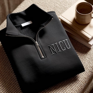 Peut inclure: Sweat-shirt noir à fermeture éclair quart de col avec les lettres brodées "NICU" en gris. Le sweat-shirt a un col montant et une fermeture éclair argentée. Le vêtement est plié sur une surface rayée.