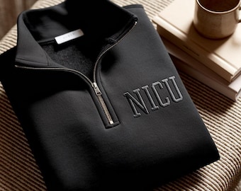 May include: Black quarter-zip sweatshirt with the embroidered letters "NICU" in gray. The sweatshirt has a stand-up collar and a silver zipper. The garment is folded on a striped surface, with a mug and books in the background.