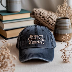 Peut inclure: Casquette de baseball noire délavée avec l'inscription brodée "Support Your Local Cemetery" en police gothique. La casquette est au centre, avec des livres, une tasse et des éléments décoratifs en arrière-plan.