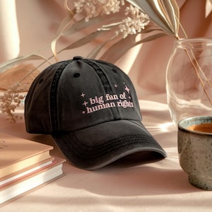 Könnte beinhalten: Schwarze Baseballkappe mit dem Text "big fan of human rights" in Rosa. Die Kappe hat einen gebogenen Schirm und ein klassisches Sechs-Panel-Design. Der Hut ist aus einem weichen, abgenutzten Material.