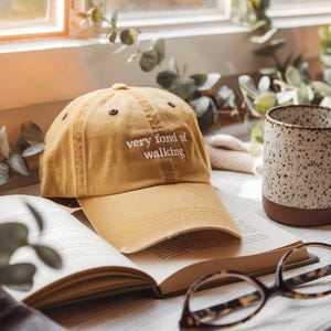 Op de afbeelding: Mosterdkleurige baseballcap met de tekst "very fond of walking" in het wit geborduurd. De pet ligt op een open boek naast een gespikkelde keramische mok en een bril. De scène speelt zich af bij een raam.