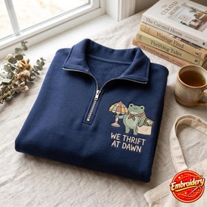 May include: A navy blue quarter-zip sweatshirt featuring an embroidered graphic of a frog holding a vintage lamp and a tote bag. Below the frog, the text reads "WE THRIFT AT DAWN" in light-colored lettering. The sweatshirt has a collar and a silver zipper.
