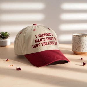 May include: A beige baseball cap with a maroon brim and top button. The cap features embroidered text in maroon that reads, "I SUPPORT A MAN'S RIGHT TO SHUT THE FUCK UP."