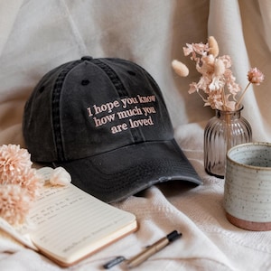 May include: A black baseball cap with the embroidered text "I hope you know how much you are loved" in pink. The cap is distressed and sits next to a notebook, pen, and small vase with dried flowers.