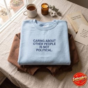 May include: A light blue crewneck sweatshirt featuring dark blue embroidered text that reads, "CARING ABOUT OTHER PEOPLE IS NOT POLITICAL." The sweatshirt is layered over a solid brown long-sleeve garment on a textured, light-colored surface.