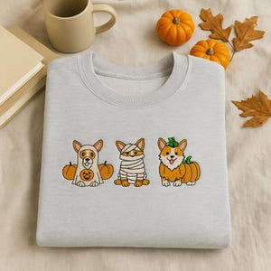 Może przedstawiać: Jasnoszara bluza z trzema haftowanymi corgi w kostiumach na Halloween. Jeden to duch, drugi mumia, a trzeci dynia. Małe dynie i jesienne liście w tle.