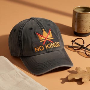 Può includere: Un cappellino da baseball grigio antracite con la scritta ricamata "NO KINGS" in oro e rosso. Il cappellino presenta un disegno a corona con una "X" rossa. Il cappello è su uno sfondo neutro con una tazza, occhiali e una foglia.