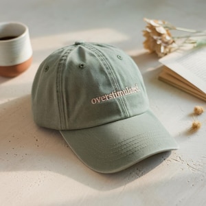 May include: A green baseball cap with the text "overstimulated" embroidered in pink on the front.