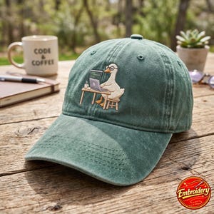 May include: A sage green baseball cap featuring an embroidered graphic of a white duck wearing round glasses, sitting on a wooden stool at a desk while working on a laptop. The design includes a small coffee mug on the desk next to the computer.