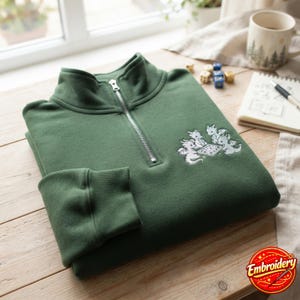 May include: A forest green quarter-zip sweatshirt featuring an embroidered design of three small dragons playing a board game on the chest. The garment has a silver-toned metal zipper pull, a high collar, and ribbed cuffs on the long sleeves.