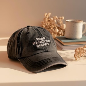 May include: A distressed black baseball cap with the text "TRUMP IS A NATIONAL DISGRACE" embroidered in white. The cap is sitting on a light-colored surface, with a mug and books in the background.