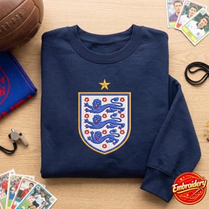 May include: A navy blue long-sleeve sweatshirt featuring an embroidered crest on the center chest. The crest displays three blue lions stacked vertically, surrounded by red roses on a white background, topped with a single gold star.
