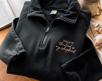 May include: Black quarter-zip sweatshirt with the text "Psalm 23 the Lord is my shepherd" in a cursive font. The sweatshirt has a collar and a silver zipper. The image is on a wooden surface with a book and a mug in the background.