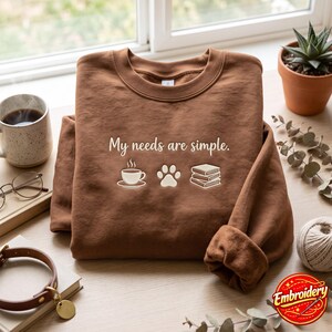 May include: A brown crewneck sweatshirt featuring the embroidered text "My needs are simple." accompanied by icons of a coffee cup, a paw print, and a stack of three books. The design is centered on the chest of the long-sleeved garment.