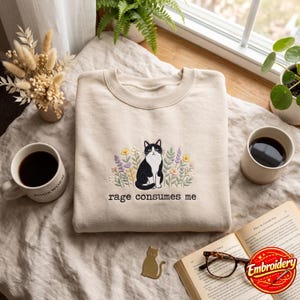 Könnte beinhalten: Ein beigefarbenes Sweatshirt mit Rundhalsausschnitt und einer Stickerei, die eine schwarz-weiße Katze inmitten bunter Blumen zeigt. Unter der Katze ist der Text "rage consumes me" in schwarzem Garn gestickt. Das Sweatshirt hat gerippte Bündchen.