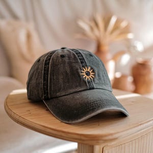 Könnte beinhalten: Eine schwarze Baseballkappe aus Denim mit einem gestickten gelben Sonnenmotiv. Die Kappe hat einen leicht abgenutzten Look.