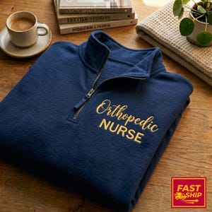 Op de afbeelding: Marineblau fleece trui met een kwart ritssluiting. De tekst "Orthopedic NURSE" is in goudkleurig garen op de voorkant geborduurd. Een kopje koffie en boeken staan op de achtergrond.