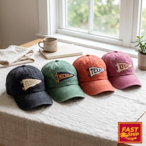 May include: Four baseball caps in charcoal, sage green, burnt orange, and dusty rose. Each cap features an embroidered pennant patch on the front with the text Florida, Michigan, Texas, or USC. The caps have a structured crown and curved brim.