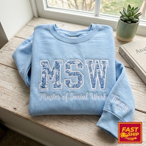 以下が含まれることがあります： 白い花柄のデザインで「MSW」の文字が刺繍された、水色のスウェットシャツ。「Master of Social Work」の文字が下に刺繍されています。袖には「Timmy」という名前とハートマークがあります。