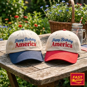 Könnte beinhalten: Zwei Baseballkappen mit beigen Kronen und gesticktem Text Happy Birthday America. Eine Kappe hat einen marineblauen Schirm, die andere einen roten Schirm. Beide Kappen haben einen Knopf an der Oberseite und gestickte Belüftungsösen.
