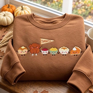 Puede incluir: Una sudadera marrón de cuello redondo con un bordado que muestra una fila de personajes de comida sonrientes tomados de la mano, incluyendo un pavo, arándanos, macarrones, puré de patatas y un trozo de tarta de calabaza con una bandera "THANKFUL".