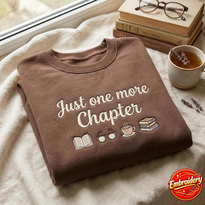 May include: A brown crewneck sweatshirt featuring the embroidered text Just one more Chapter in a cream color. Below the text are small embroidered icons of an open book, a pair of glasses, a steaming cup of coffee, and a stack of three books.