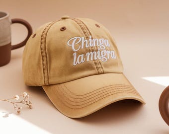 May include: A tan baseball cap with the phrase "Chinga la migra" embroidered in white script. The cap has a curved brim and a classic six-panel design. The hat is made of a durable fabric and features a brown button on top.