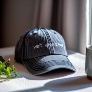 May include: A gray baseball cap with the text "wait, i see a rock" in white. The cap has a curved brim and a stitched design. The hat is made of a soft material and is a casual accessory.